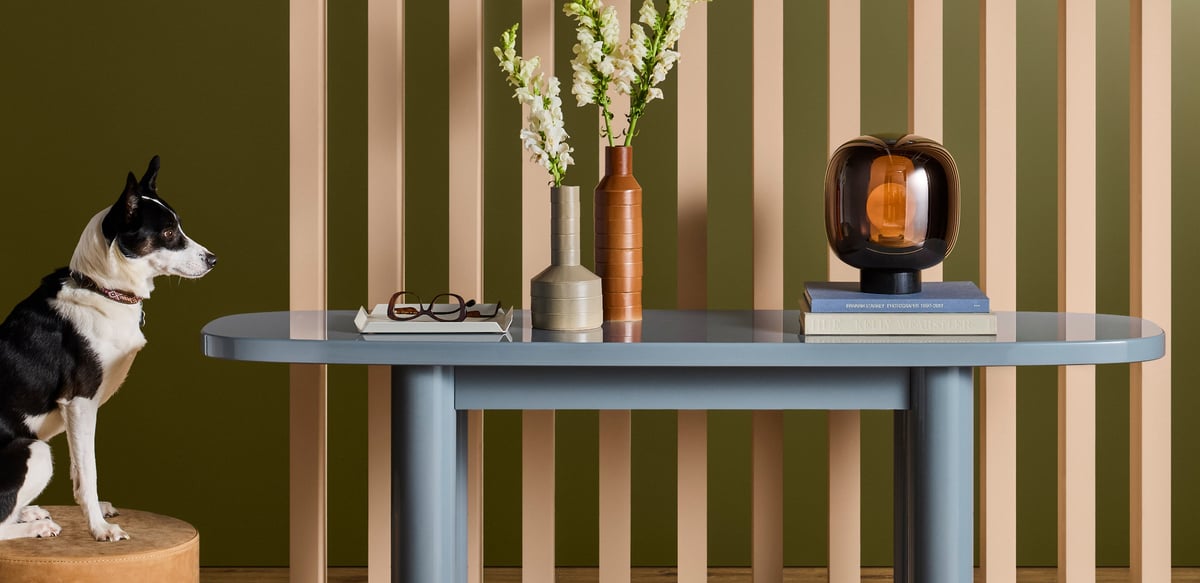 A dog perched on a tan stool beside a blue-gray Blu Dot table with sculptural vases