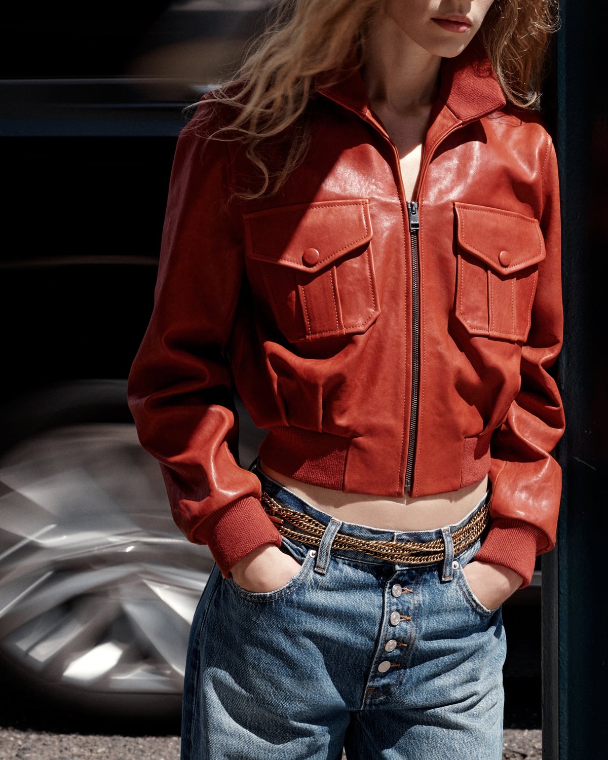 A model wearing a red leather jacket and blue jeans beside a car