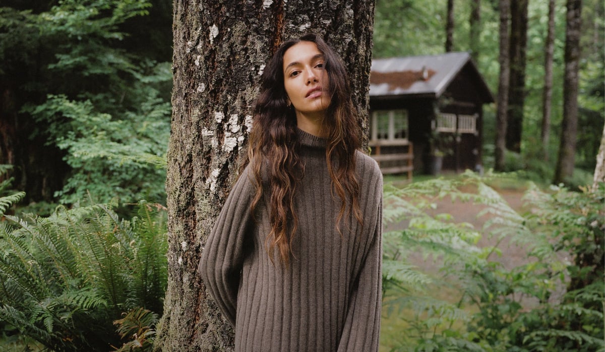 A model in a brown ribbed sweater standing beside a tree in a wooded setting