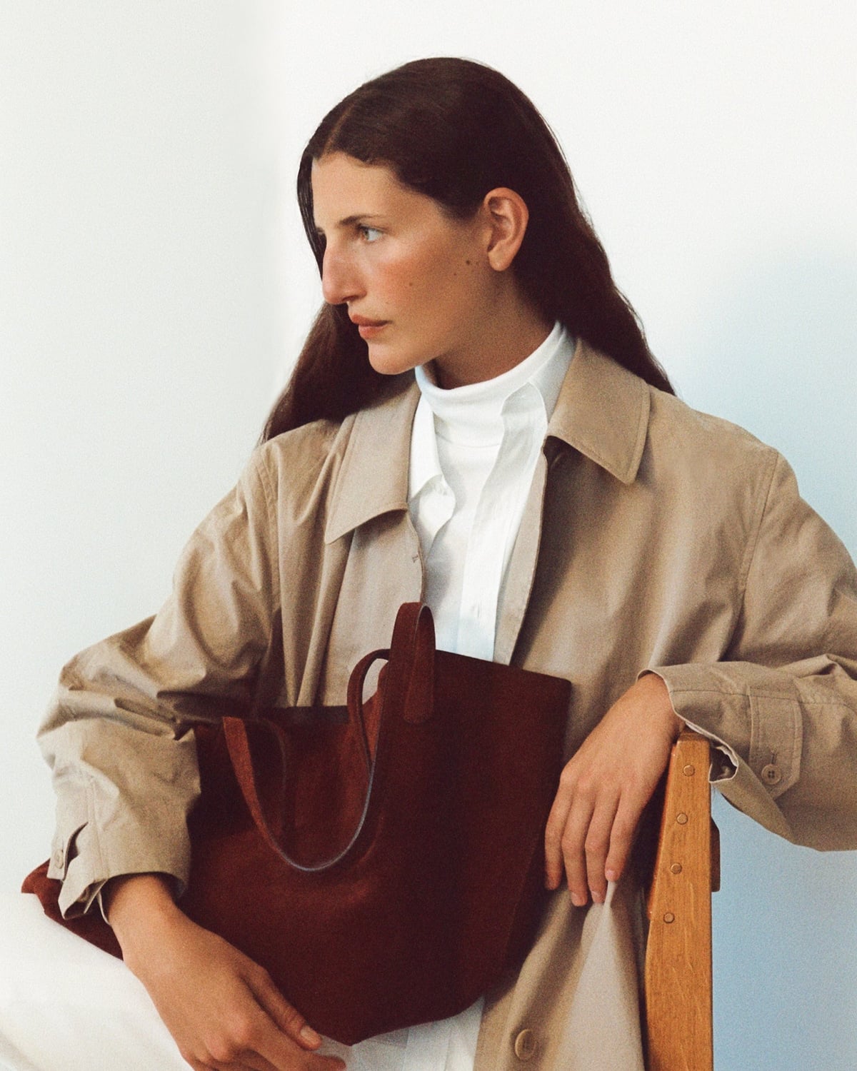 A seated model holding a burgundy handbag and wearing a beige coat