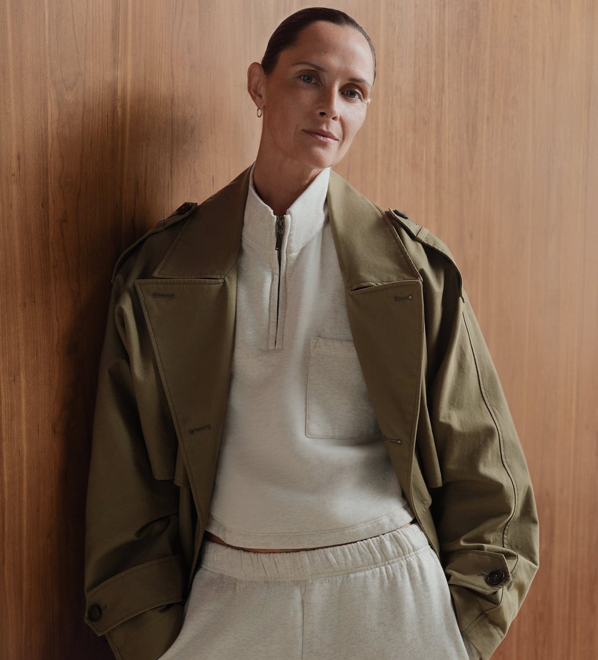 A MATE model wearing a cream half-zip and matching sweatpants beneath an olive jacket against a wood-paneled wall