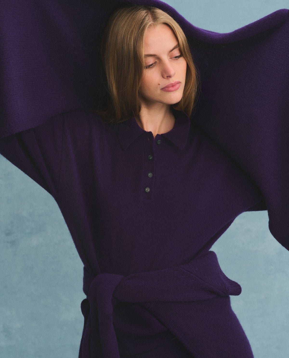 A model wearing a plum knit sweater with raised arms against a purple background